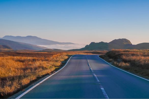Lonely country road winding through a vast hilly landscape, with mountains in the background and golden sunlight bathing the scene.